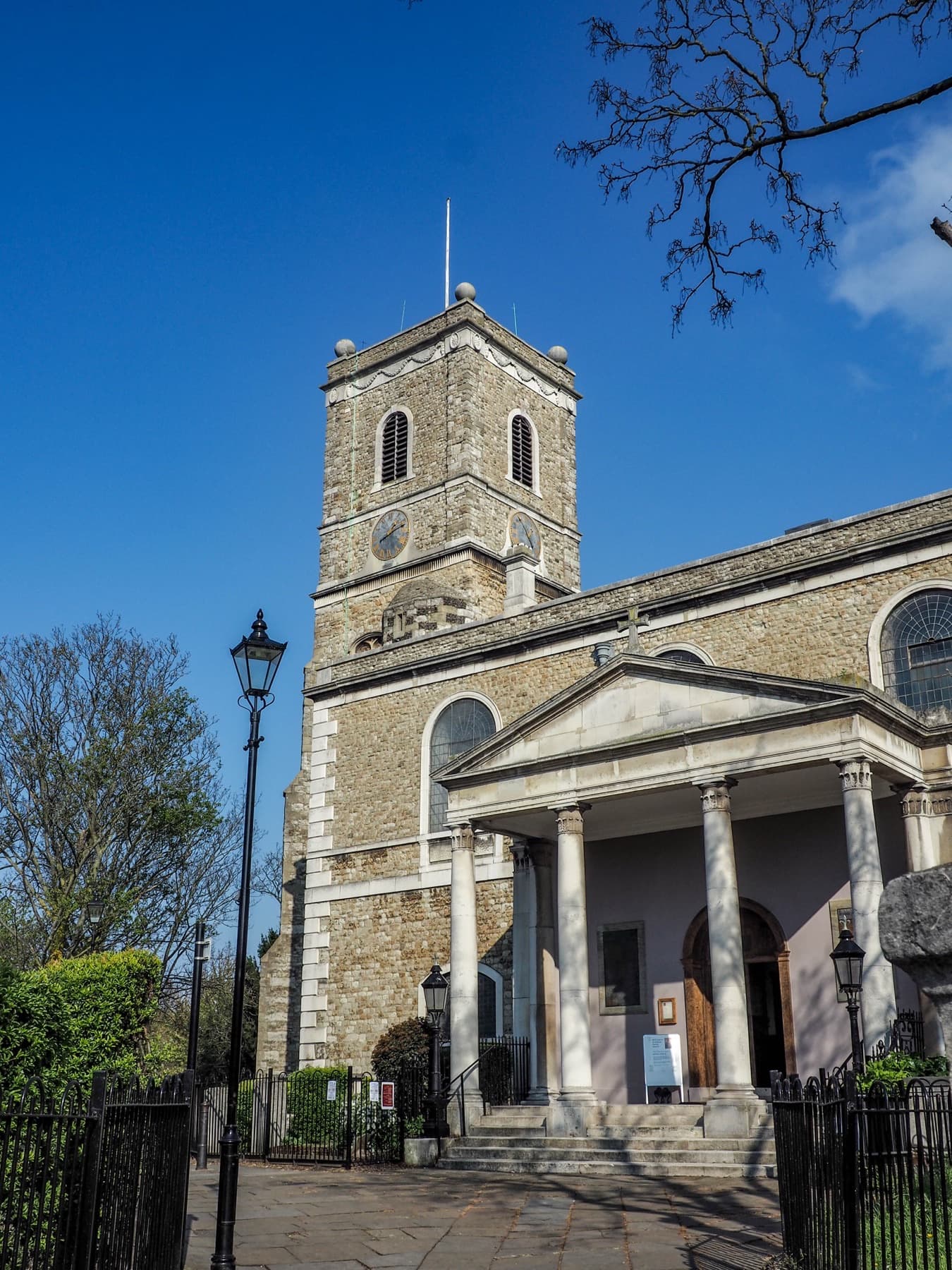 The west front of St Mary the Virgin from the churchyard, its portico and columns in sunlight, with the tower rising behind.